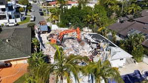 Orange excavator demolishing a house between neighboring homes with a debris truck nearby - cypress line services - demolition in louisiana