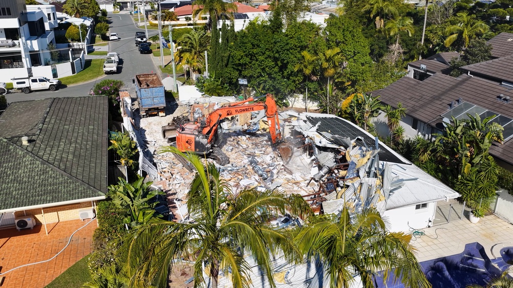 Orange excavator demolishing a house between neighboring homes with a debris truck nearby - cypress line services - demolition in louisiana